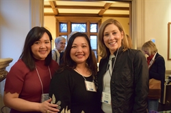 [ai] Three women smiling together at an event, standing in a well-lit room with large windows. The woman on the left is wearing a maroon top, the center woman has a friendly expression in a black sweater, and the woman on the right is dressed in a black jacket.